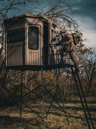 A man wearing hunting gear, carrying a bow, climbing the ladder on his Grizzly Hunting Blind Tower Kit, to his Grizzly Alpha Combo Hunting Blind. The trees are mostly bare, with some green grass covered in leaves below, suggesting that it is autumn. 