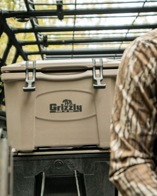 A tan Grizzly Coldware cooler with black latches and a black logo, shown on a vehicle rack with a person in camouflage clothing partially visible on the right.