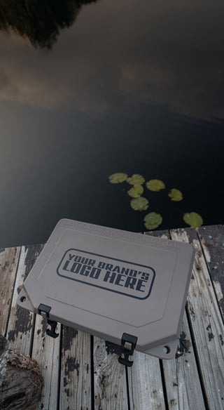 The image shows a hard side Grizzly cooler with an imprint on the top of the lid that reads "Your brand's logo here". The cooler is tan with the custom branding text in black. There cooler is on a dock. You can see lily pads in the water behind the cooler. Custom branding for personal or corporate gifts is available from Grizzly Coolers.