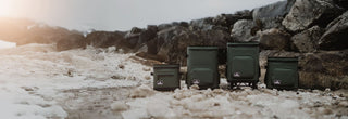 Four soft side coolers are lined up in the image along a background of large rocks and sitting on the snowy ground. The soft coolers are the new soft portable coolers from Grizzly Coolers. The soft coolers are all in the color lunar green. On the ends are the smaller soft side coolers, the Drifter 8 and Drifter 16. In the middle are the larger capacity portable coolers, the new backpack cooler and the Drifter 32 is a soft side cooler on wheels. 