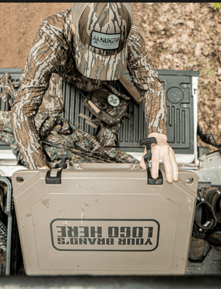 The image is an aerial view of the lid of a Grizzly cooler that someone is unlatching and opening. On top of the lid, you can see an imprint that reads "your brand's logo here". Grizzly Coolers offers custom branding on their products including custom branded coolers and custom branded drinkware. The hard sided cooler in this image is tan and is in a truck bed with the tailgate down. There's a turkey decoy and hunting boots in the image on each side of the cooler.