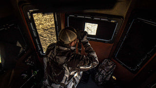 The image shows a man wearing camouflage clothing and a trucker hat, sitting inside the Alpha Hunting Blind. The man is using binoculars, looking out of the peephole of the hunting blind window covered with a blackout shade. The blackout shades keep you completely hidden by ensuring that no light gets in or out. The middle peep window of the blackout shade has an easy-to-operate magnetic open/closure for viewing and ranging with one-handed, noise-free motion.