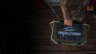 The image shows a man's hand carrying a hard side Grizzy cooler by the handle where you can see the top of the lid with the words "Team Realtree" printed over the pattern of Realtree camouflage. The Grizzly hard sided cooler is in the color Ranger Green and the man is also wearing Realtree camouflage printed shirt and pants. The floor in the background is wood.