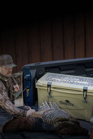 The image shows a man wearing Realtree camouflage holding a large turkey by the leg on the back of his tailgate. The turkey is laying in front of a hard side Grizzly cooler with the words Team Realtree printed on the top of the lid. The background of the lid is in Realtree camouflage and the Grizzly logo on the front of the cooler is also printed in Realtree camouflage. The cooler is Ranger Green in color.