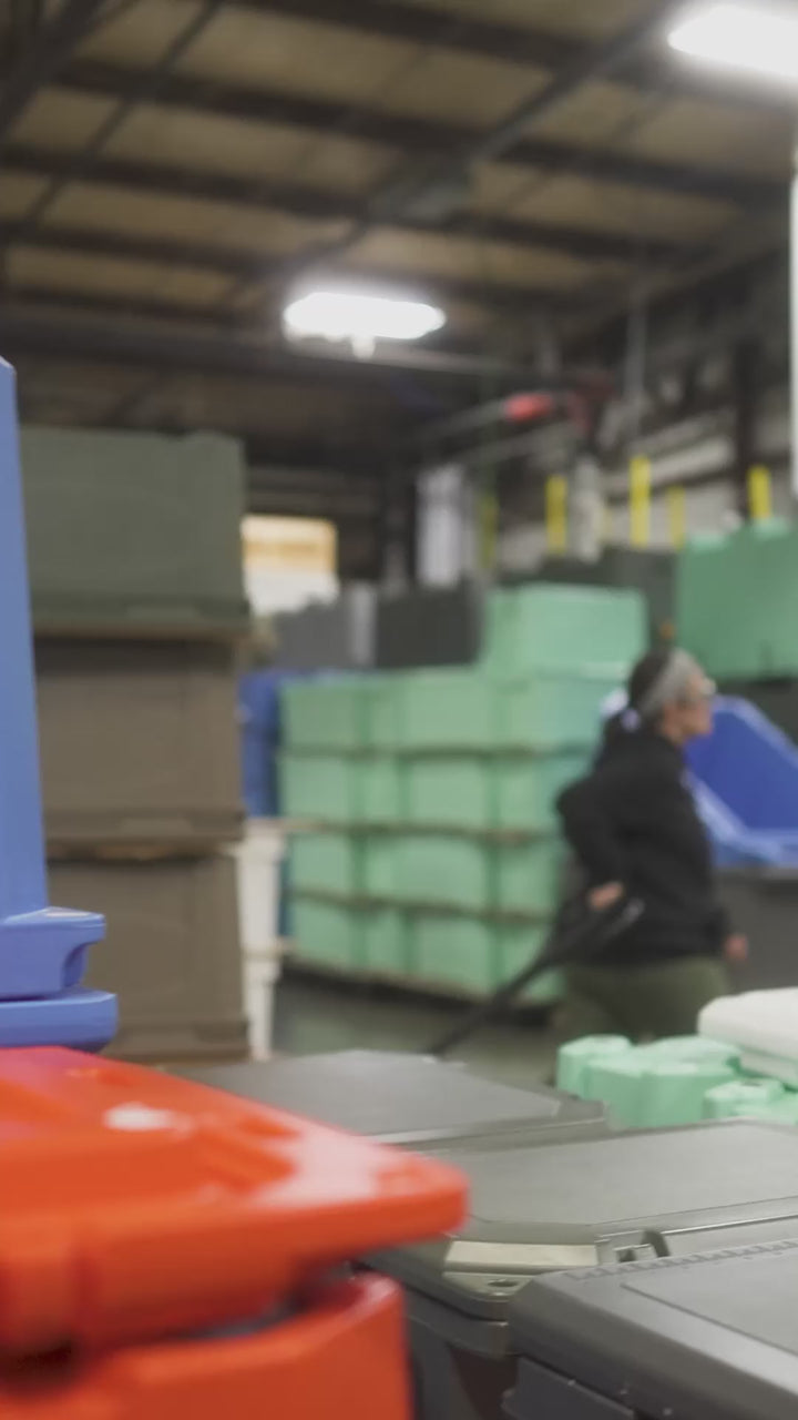 Grizzly Coolers warehouse with stacks of coolers in various colors. A female worker is shown in the warehouse wearing safety glasses while pulling a cart.