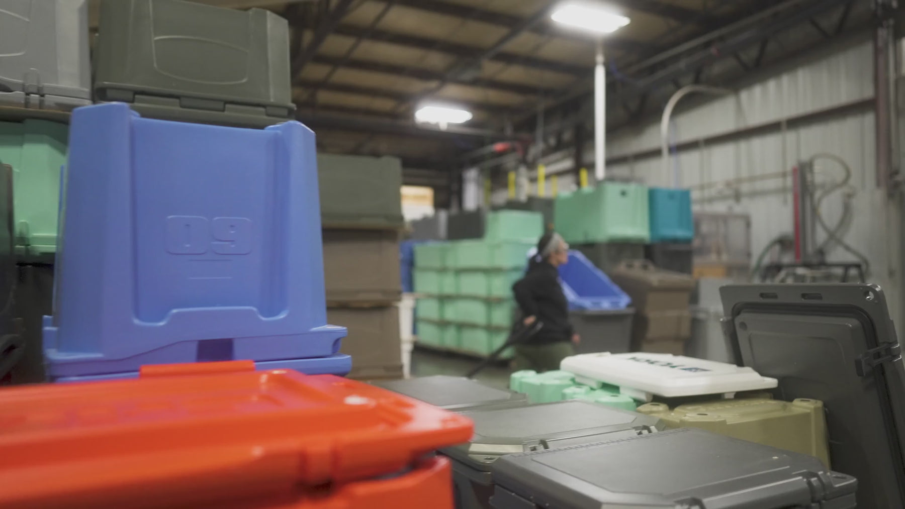 Grizzly Coolers warehouse with stacks of coolers in various colors. A female worker is shown in the warehouse wearing safety glasses while pulling a cart.