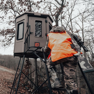 The image shows a man wearing camouflage and an orange vest, climbing the ladder to his Grizzly Alpha Hunting Blind. The man has a rifle on his back and there is a tree with a few brown leaves left on it in the background as it is whitetail deer hunting season. The hunting blind is a brown color which makes it blend in well with the surroundings.