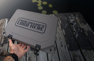 The image shows a hard side Grizzly cooler with an imprint on the top of the lid that reads "Your brand's logo here". The cooler is tan with the custom branding text in black. There cooler is on a dock. You can see lily pads in the water behind the cooler. Custom branding for personal or corporate gifts is available from Grizzly Coolers.